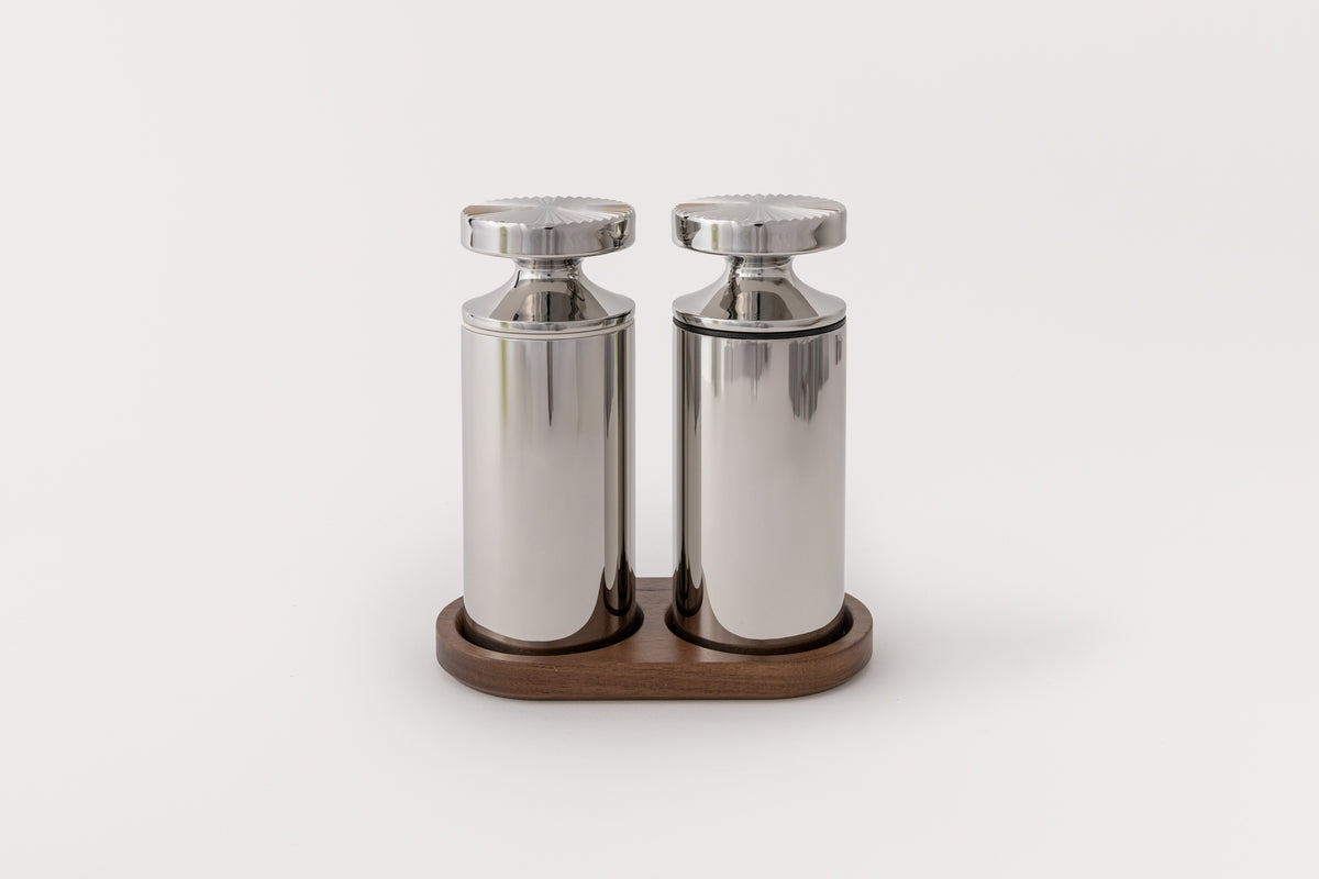 Two stainless steel grinders with one featuring a black band and the other featuring a white band, placed on a walnut stand.