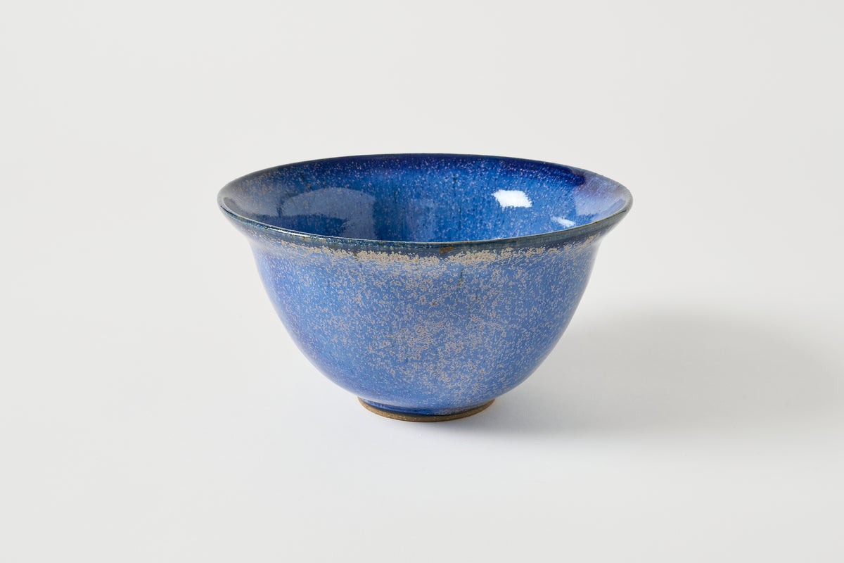 A blue ceramic bowl with a speckled glaze and a small footed base, photographed on a clean light gray background.