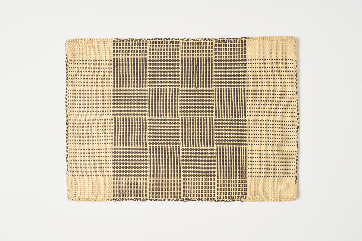 A rectangular woven placemat with a geometric pattern, featuring a central basketweave motif in black on a natural beige background, set against a plain white studio backdrop.