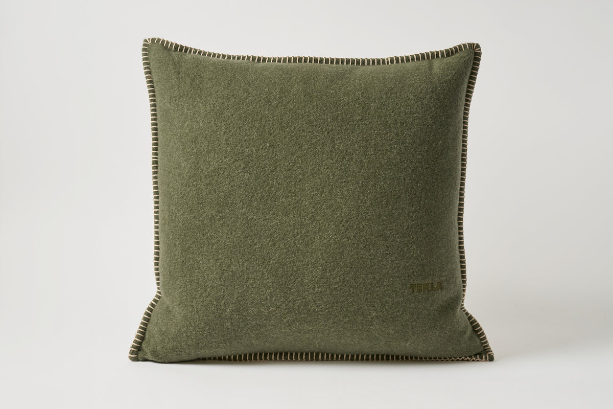 An olive green textured throw pillow with dark brown contrast stitching and a small gold 'TULLA' logo on the bottom right, set against a plain light gray background.