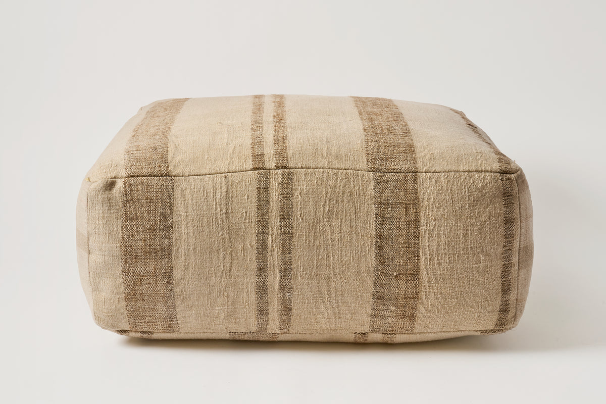 A square floor pouf or ottoman upholstered in a textured woven fabric with vertical stripes in beige and brown, shown against a plain light gray background.