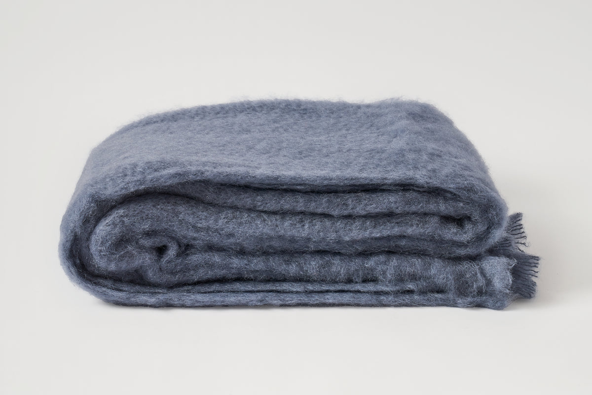 A folded dusty blue wool blanket with fringed edges, displayed on a clean off-white background.