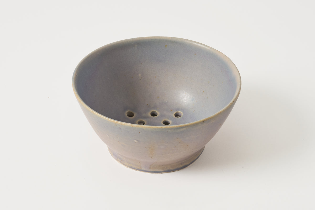 A small ceramic bowl with a perforated bottom, featuring a matte slate-blue to gray glaze with subtle earthy brown speckles near the rim, set against a plain white background.