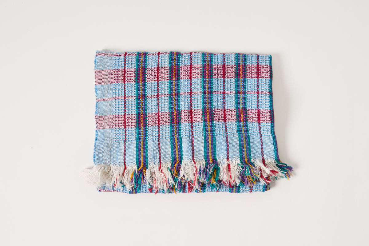 A folded woven textile with a plaid pattern in light blue, red, and dark blue, finished with fringed edges, shown on a plain white background.