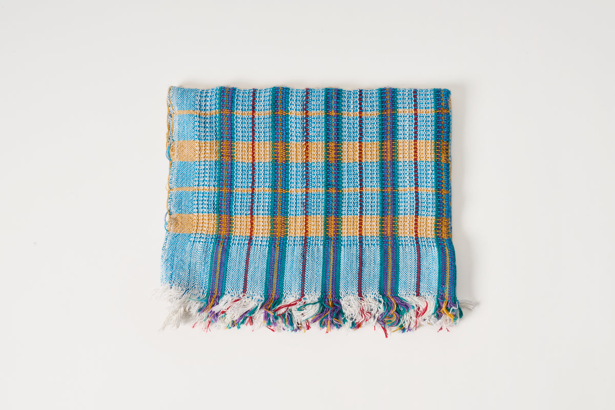 A square woven textile with fringed edges, featuring a plaid pattern in light blue, mustard yellow, and red on a white background.