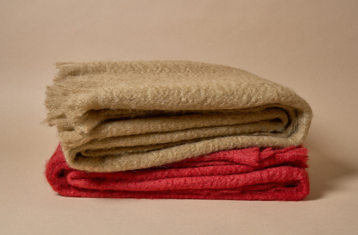 Two folded textured woolen blankets, one in natural beige and one in red, stacked on a warm beige background.
