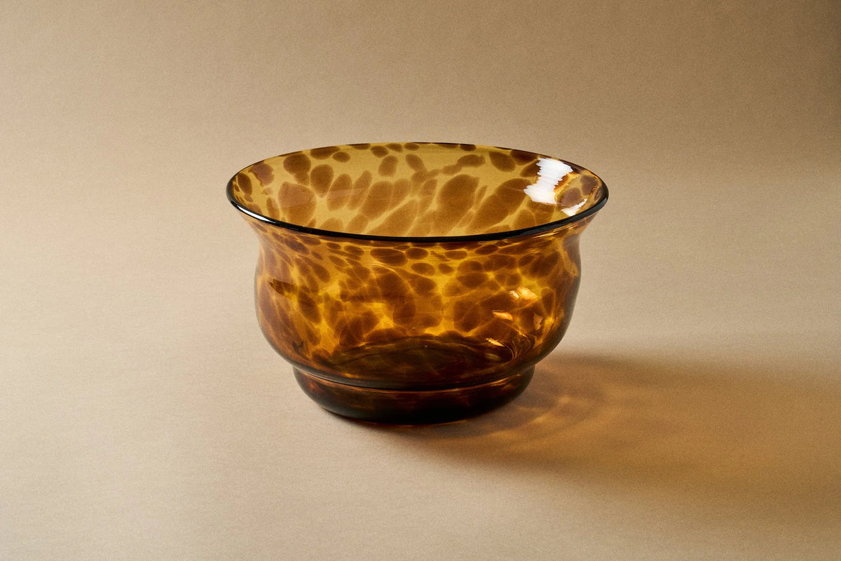 A small amber glass bowl with a mottled leopard-like pattern of darker brown and golden yellow, set against a solid beige background.