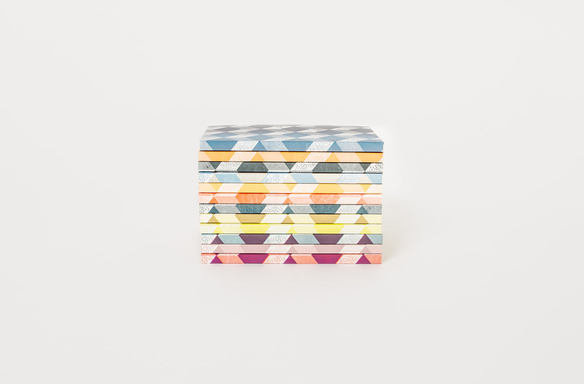 Stack of Astier de Villatte notebooks with multicolor geometric patterns on the covers, placed on a white surface.