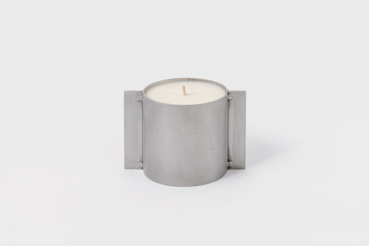 A cylindrical soy wax candle with a metallic silver finish and an organic cotton wick, designed by FDK Floral in collaboration with John Wells.
