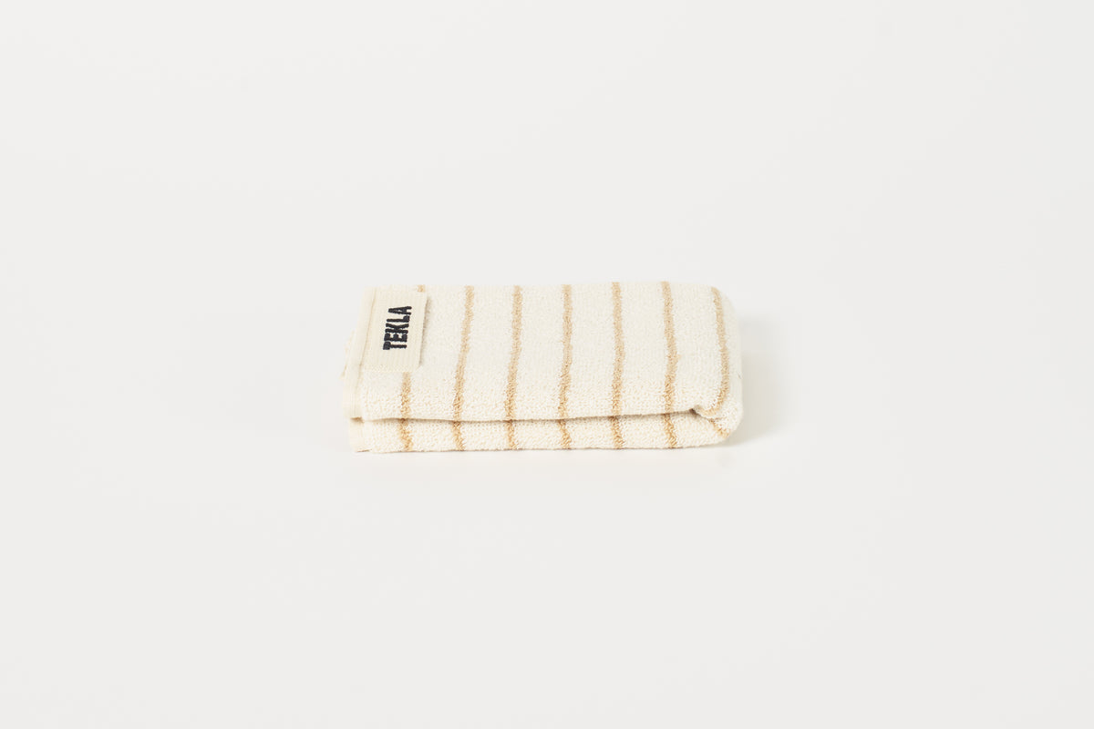 A folded beige and white striped washcloth on a white background with the brand name 'Tekla' visible