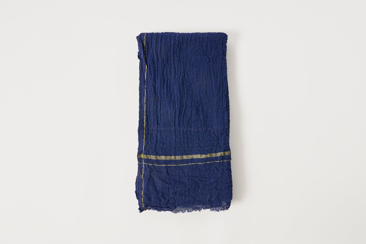 A folded dark blue crinkled fabric scarf with thin gold stripes near the bottom edge, displayed against a clean off-white background.