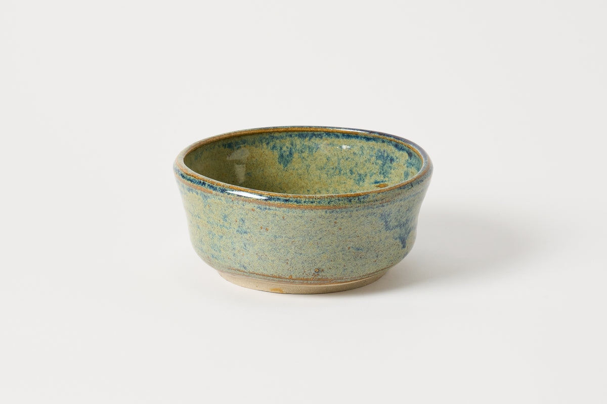 A small handmade ceramic bowl with a textured, speckled glaze in shades of sage green, blue, and beige, shown on a clean white background.