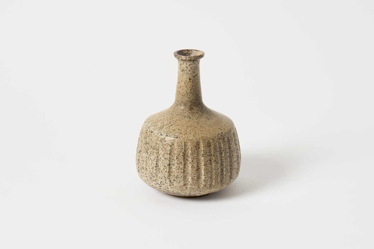 A small ceramic vase with a rounded ribbed body and a narrow neck, featuring a speckled beige and brown glaze, set against a plain light gray background.