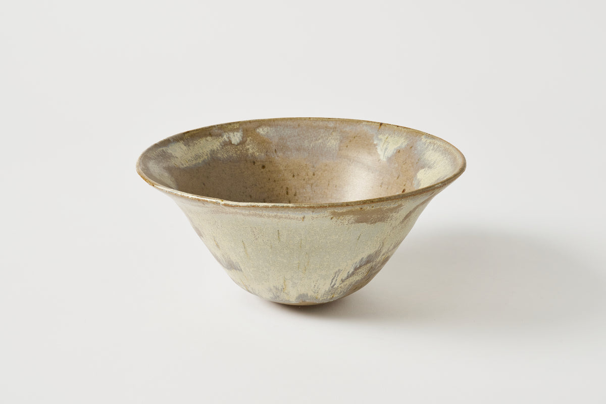 A handmade ceramic bowl with a wide, flared rim and a tapered, conical body, featuring a textured glaze with a mix of beige, brown, and gray tones, set against a plain light gray background.