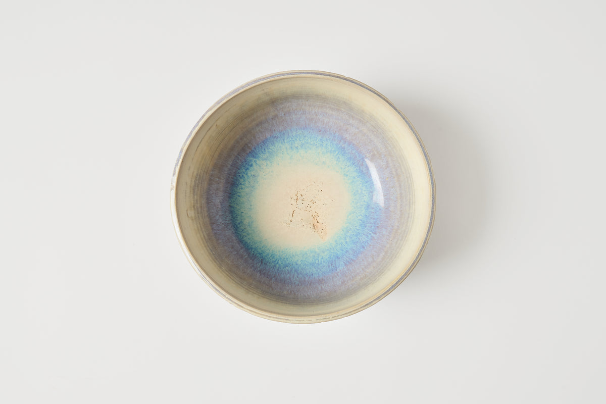 A handmade ceramic bowl viewed from directly above, featuring a glazed interior with a gradient of blue, purple, and cream, surrounded by a natural unglazed beige rim, set against a plain light gray background.