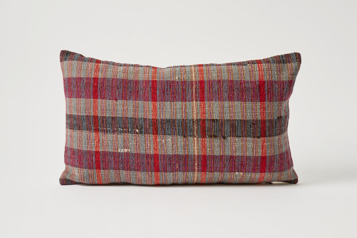 A rectangular throw pillow with a rustic plaid pattern in red, brown, beige, and gray, photographed against a neutral light gray background.