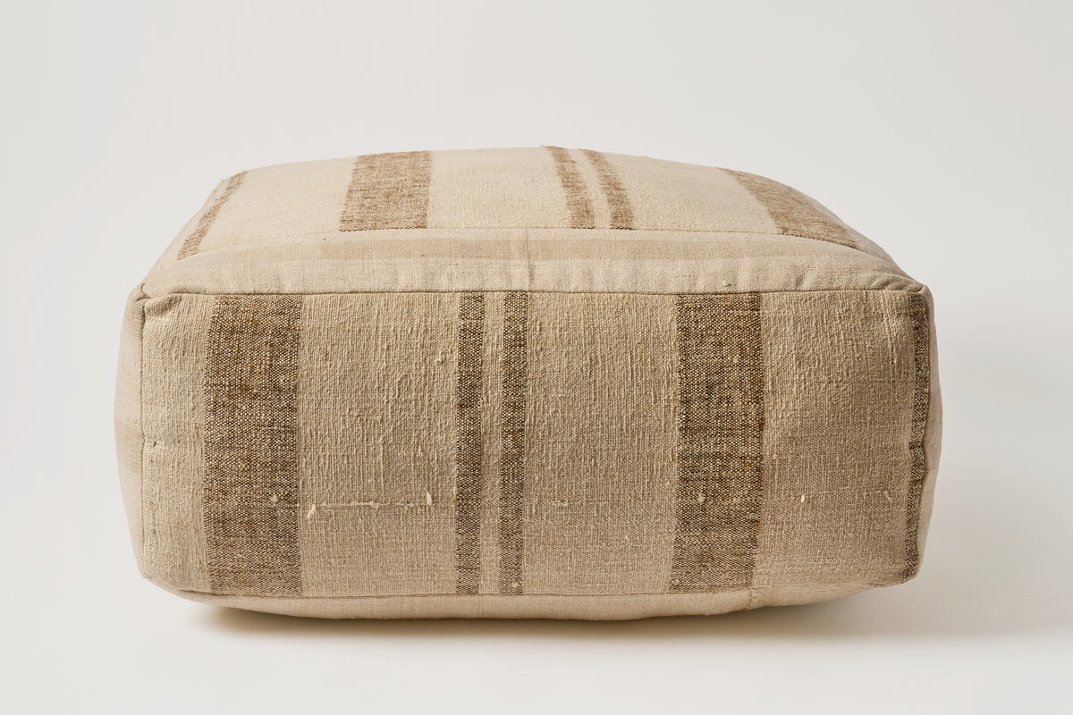 A square floor pouf or ottoman with a textured woven fabric in beige and brown vertical stripes, photographed against a neutral light gray background.