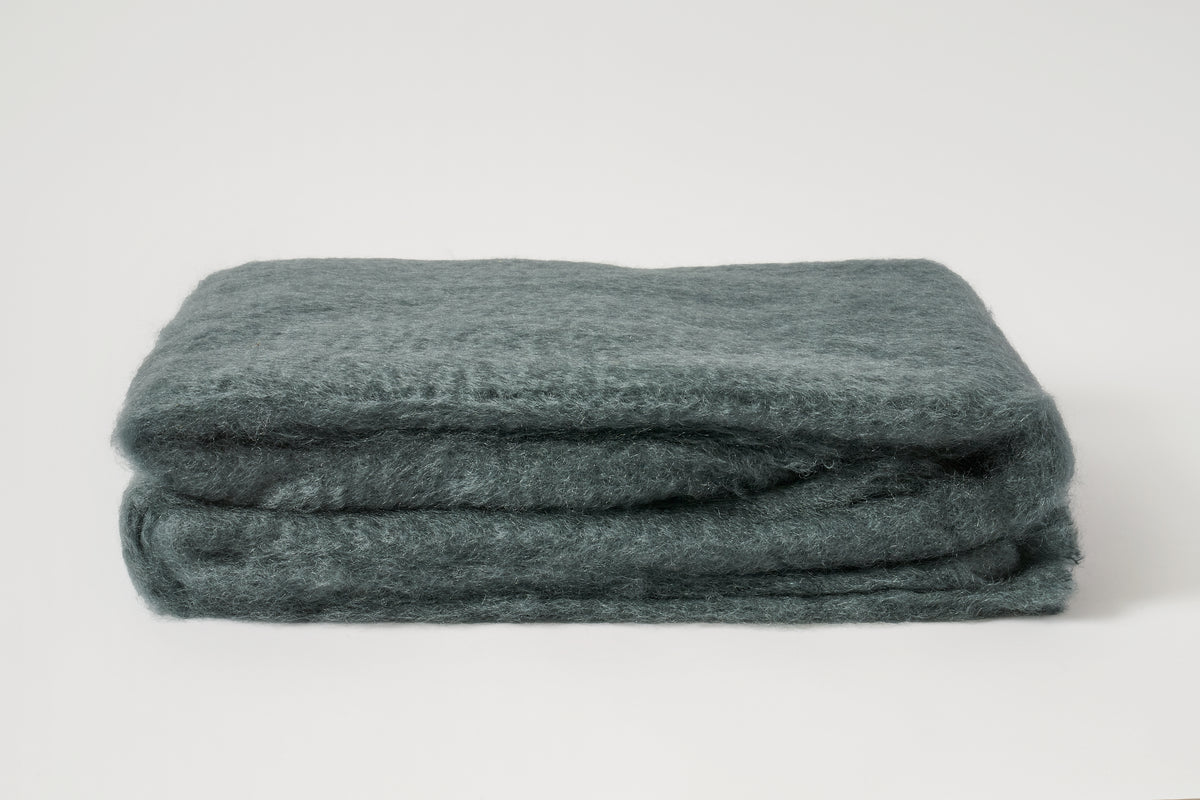A folded, thick textured wool blanket in a muted slate blue-gray color, photographed on a seamless light gray background.