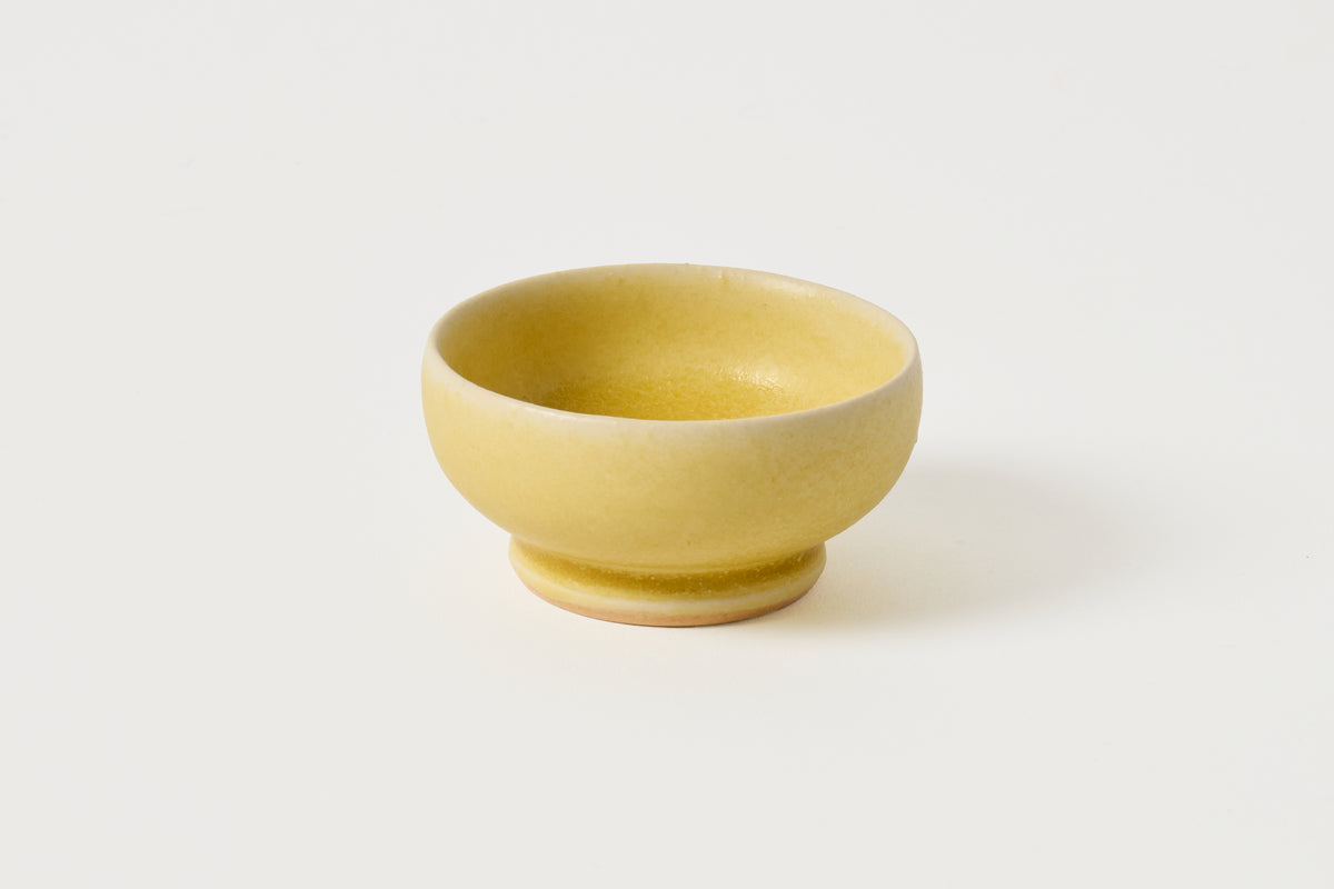 A small, round ceramic bowl with a textured matte yellow glaze and a matching footed base, photographed on a clean, solid off-white background.