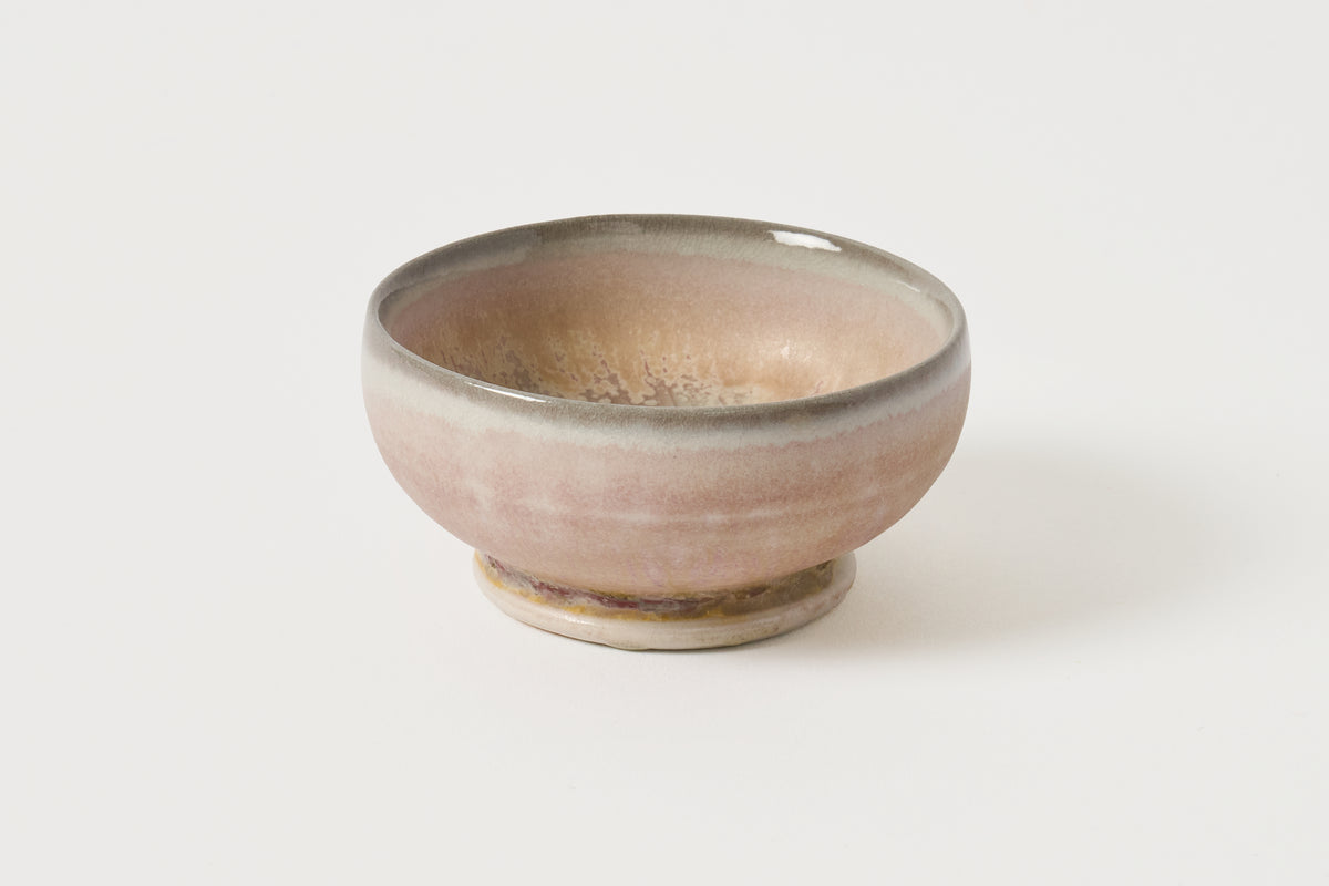 A small handmade ceramic bowl with a dusty pink and beige glaze and a dark rim, set against a plain white background.