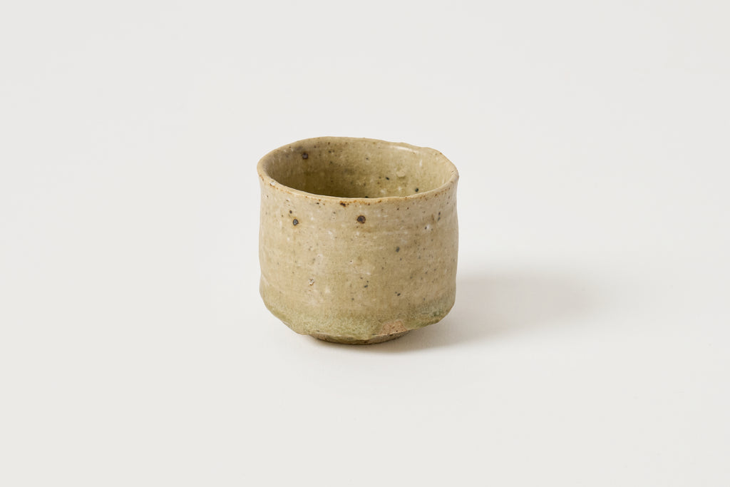A small handmade ceramic cup with a rustic, slightly tapered shape, featuring a speckled sage green and beige glaze on a clean white background.