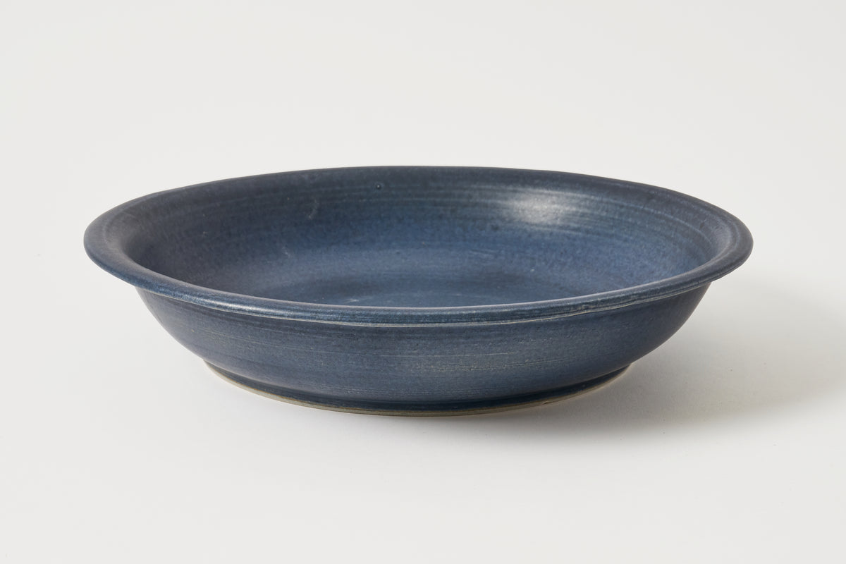 A round ceramic shallow bowl with a textured, matte dark blue glaze, photographed on a clean white background.