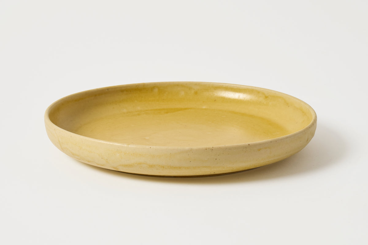 A shallow round ceramic plate in a warm mustard yellow glaze, photographed on a clean white background.