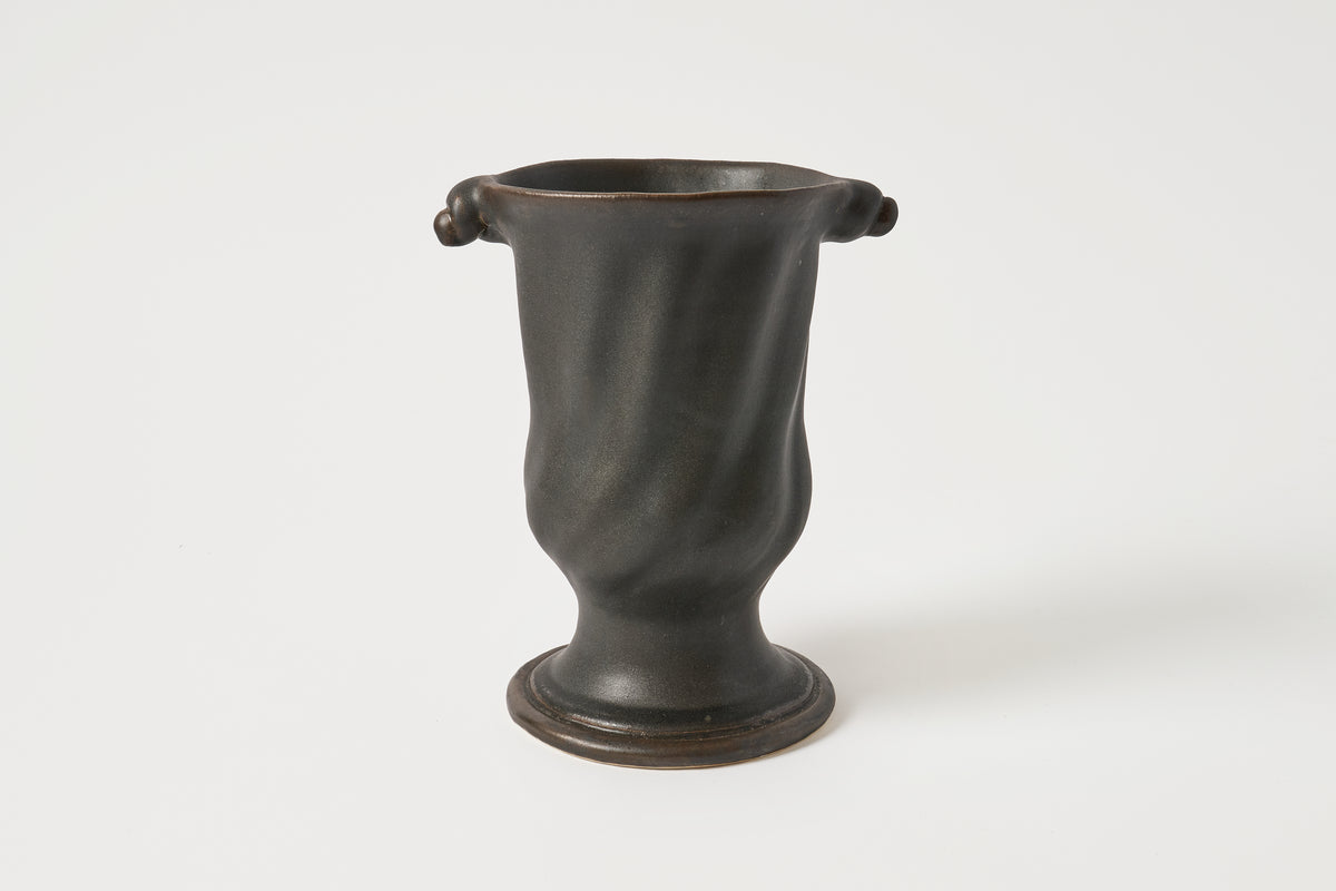 A dark, matte-finished ceramic vase with a flared base and two small loop handles near the rim, photographed against a plain light gray studio background.