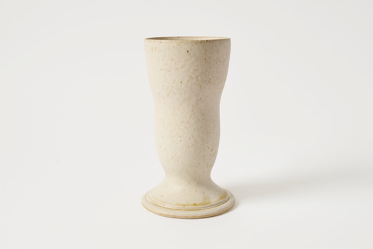 A small ceramic cup or tumbler with a flared base, made from a speckled cream-colored clay, shown on a plain white background.