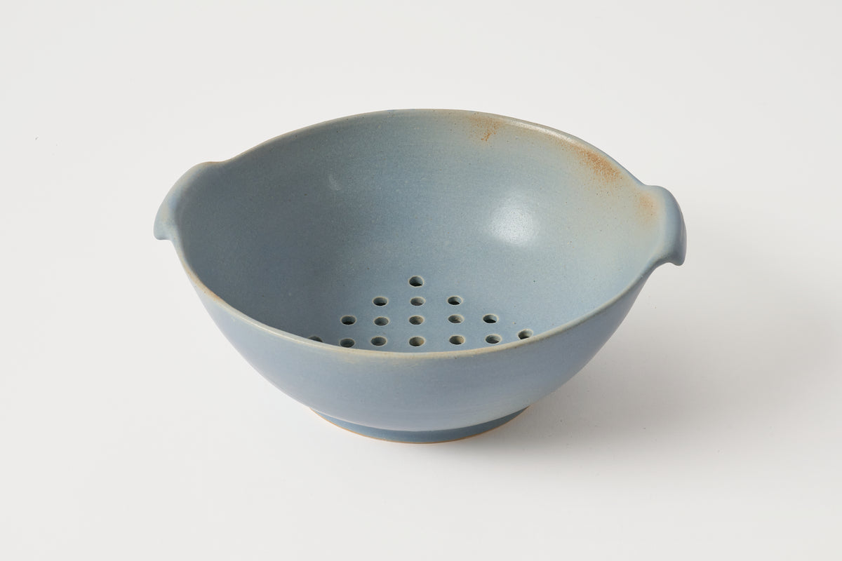 A light blue ceramic colander bowl with side handles and a perforated bottom, shown on a clean white background.