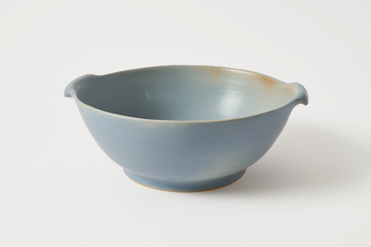 A light blue ceramic bowl with small side handles and a subtle warm brown glaze along the rim and base, photographed on a clean white background.