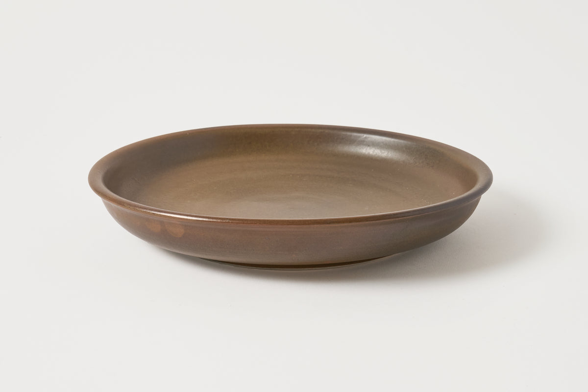 A round ceramic shallow bowl or plate with a matte brown glaze, photographed on a clean white background.