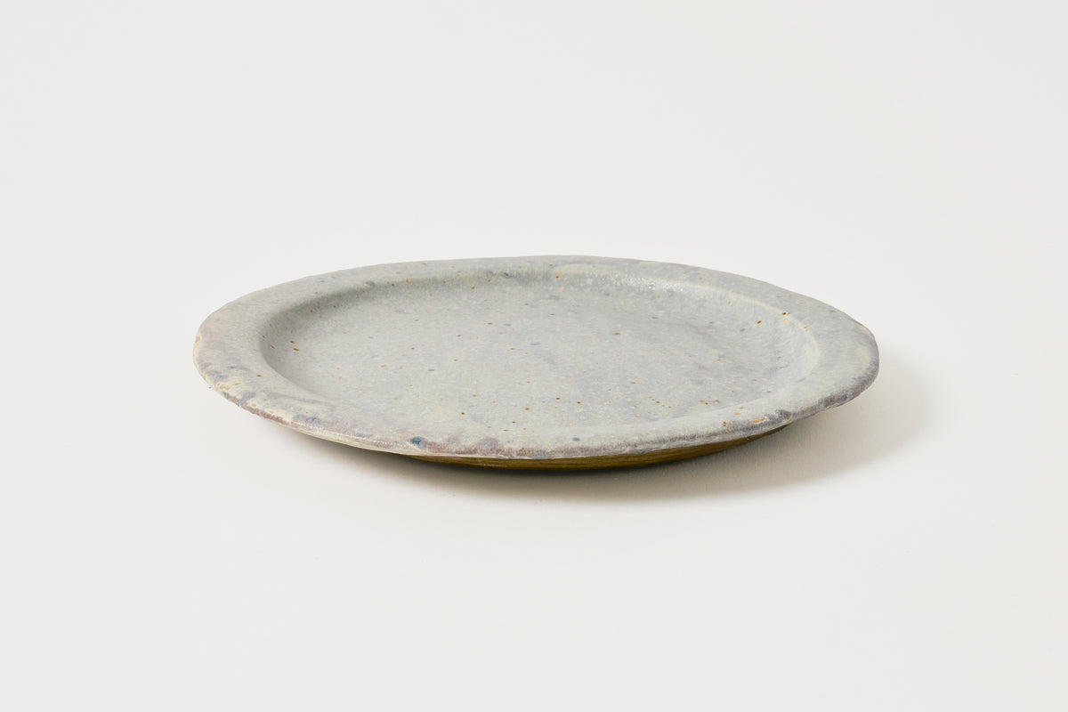 A small round ceramic plate with a textured, speckled light gray-blue glaze and a subtle warm yellow-brown edge, photographed on a clean white background.