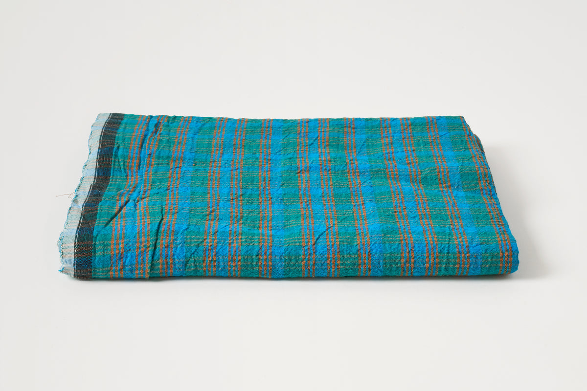 A folded woven textile, likely a scarf, shawl, or small blanket, featuring a vibrant blue and teal plaid pattern with orange stripes and a dark border, set against a plain white background.