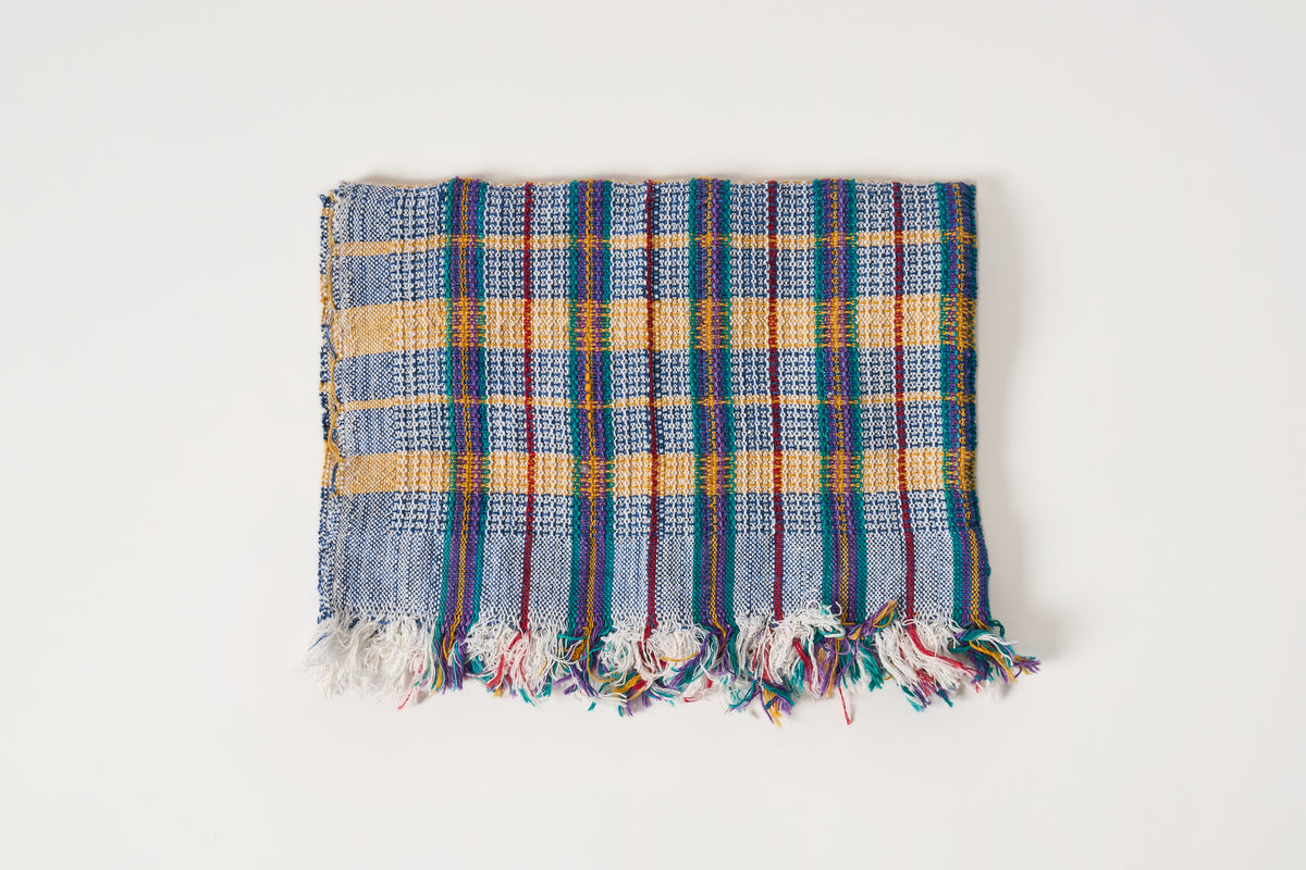 A square woven towel with a blue, yellow, and red plaid pattern and fringed edges, displayed flat against a plain white background.