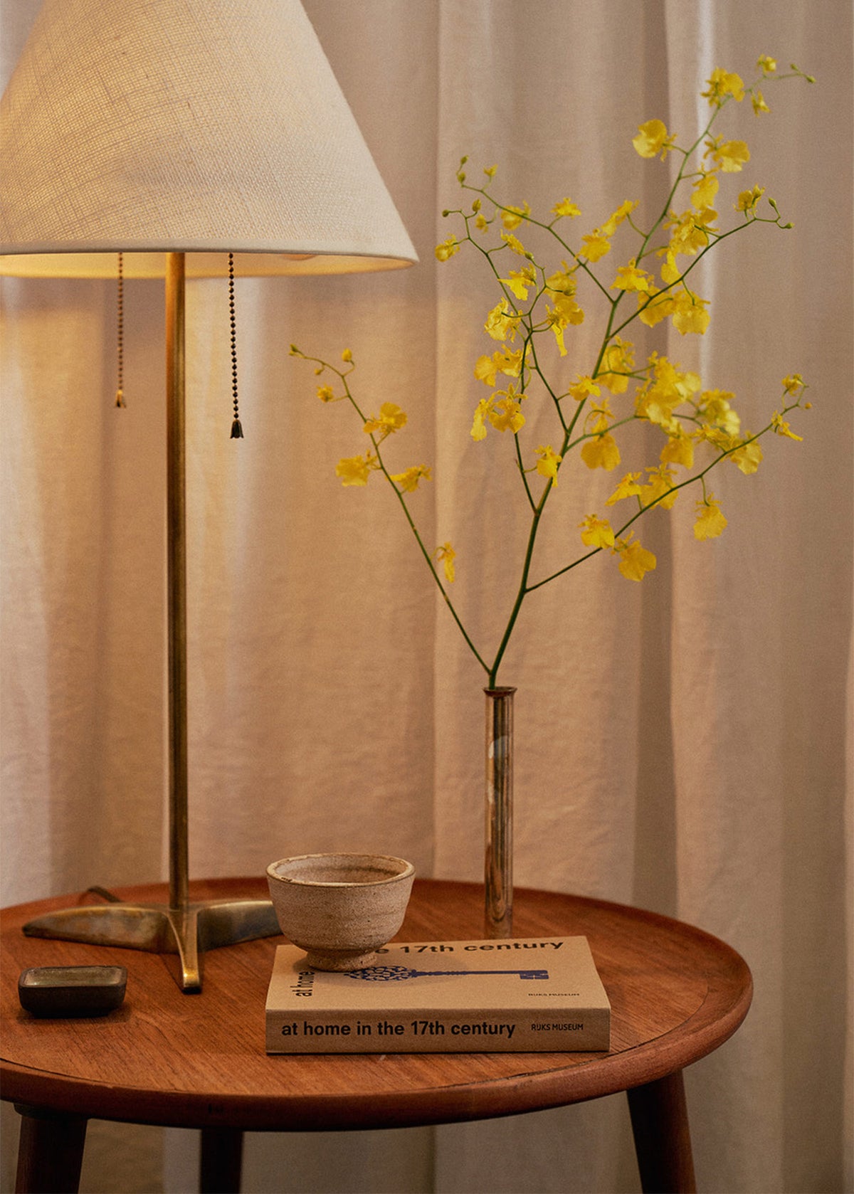 A warm, styled interior scene featuring a brass table lamp with a beige fabric shade next to a tall, slender vase holding yellow flowering branches, set on a round wooden side table with a small ceramic bowl and a book.