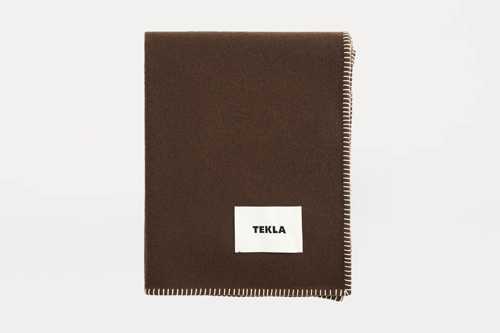 A dark brown woven throw blanket with cream-colored fringed edges and a white square label reading 'TEKLA' in black text, shown on a plain light gray background.
