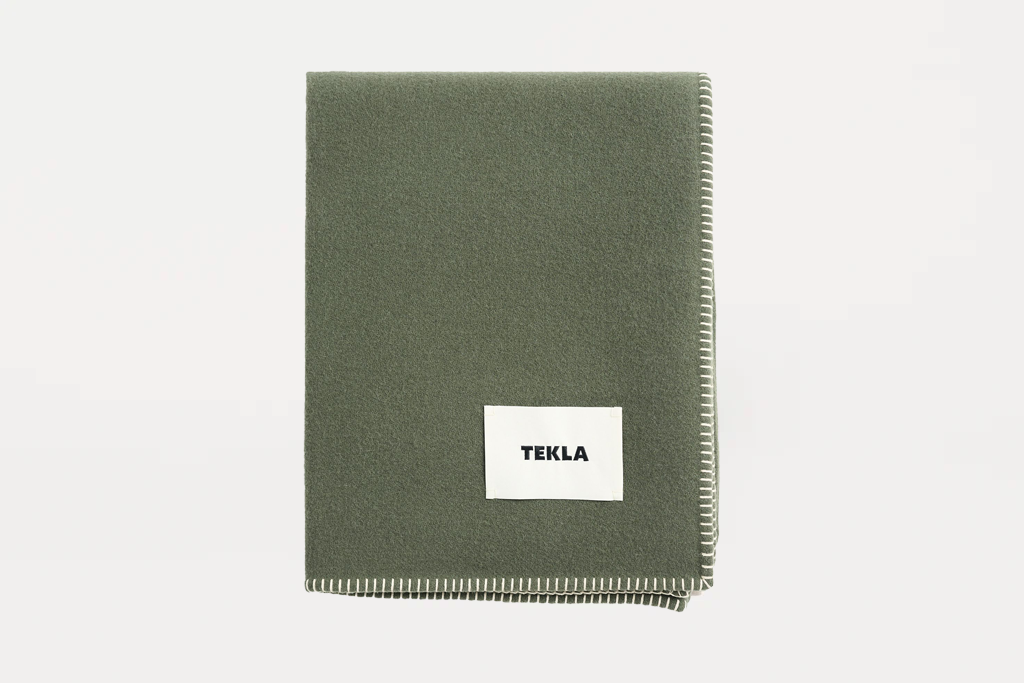 An olive green woven throw blanket with a white rectangular label reading 'TEKLA' in black text, displayed flat against a clean white background.